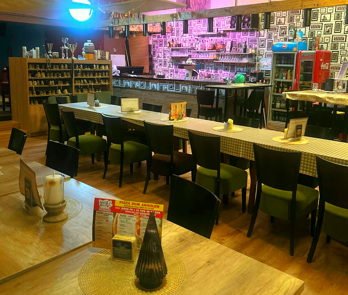 Das Restaurant mit langen Tischen und grünen Stühlen, dahinter die Bar